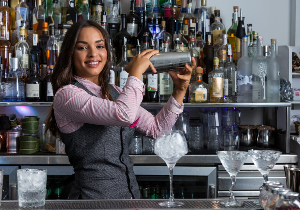 Happy young ethnic female barkeeper mixing drinks in shaker while preparing cocktail with ice cubes at bar counter